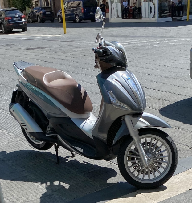 Ein Motorroller mit einem Helm darauf auf einer Straße geparkt, mit Gebäuden, Bäumen, Pfählen, Menschen und Outdoor-Möbeln im Hintergrund.