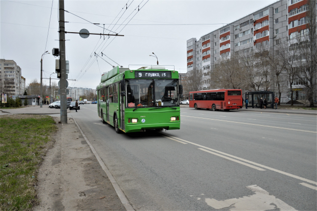 Ein grüner Bus fährt auf einer Straße mit hohen Gebäuden, Gras auf der linken Seite, Strommasten mit Drähten, Bäumen im Hintergrund und einem klaren blauen Himmel.