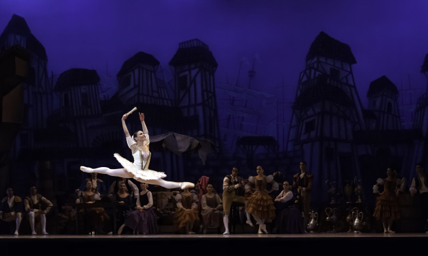 Eine Balletttänzerin in einem weißen Tutu und Spitzenschuhen tanzt auf der Bühne, während das Publikum zusieht, mit einem Schlossgemälde im Hintergrund.