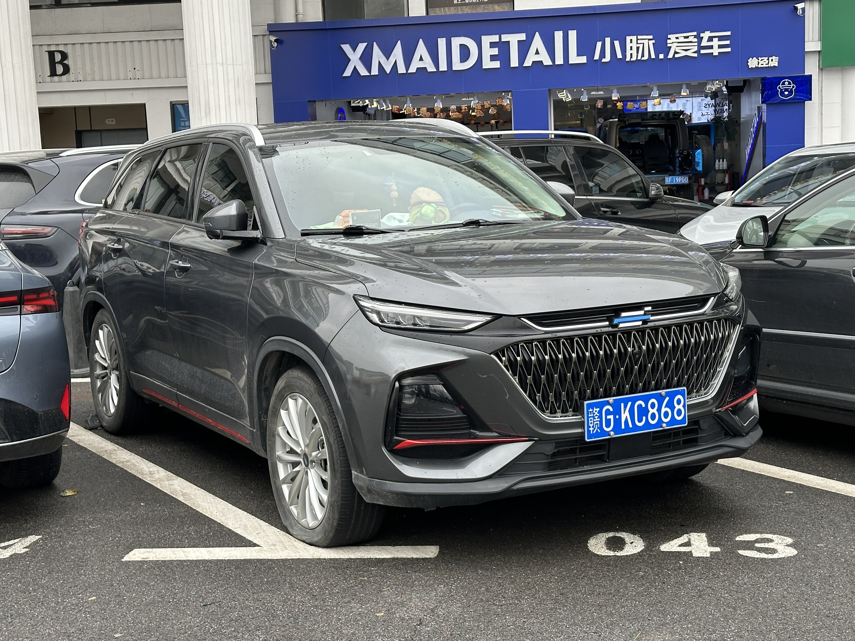 Graues Auto vor einem Gebäude mit Säulen und einem Namensschild in China geparkt, mit Menschen und Gegenständen im Inneren sichtbar.
