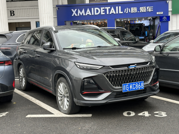 Graues Auto vor einem Gebäude mit Säulen und einem Namensschild in China geparkt, mit Menschen und Gegenständen im Inneren sichtbar.
