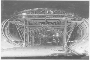 Ein Schwarz-Weiß-Foto eines im Bau befindlichen Tunnels mit Eisenstangen und Lampen, die den Bereich beleuchten, und Text am unteren Rand.