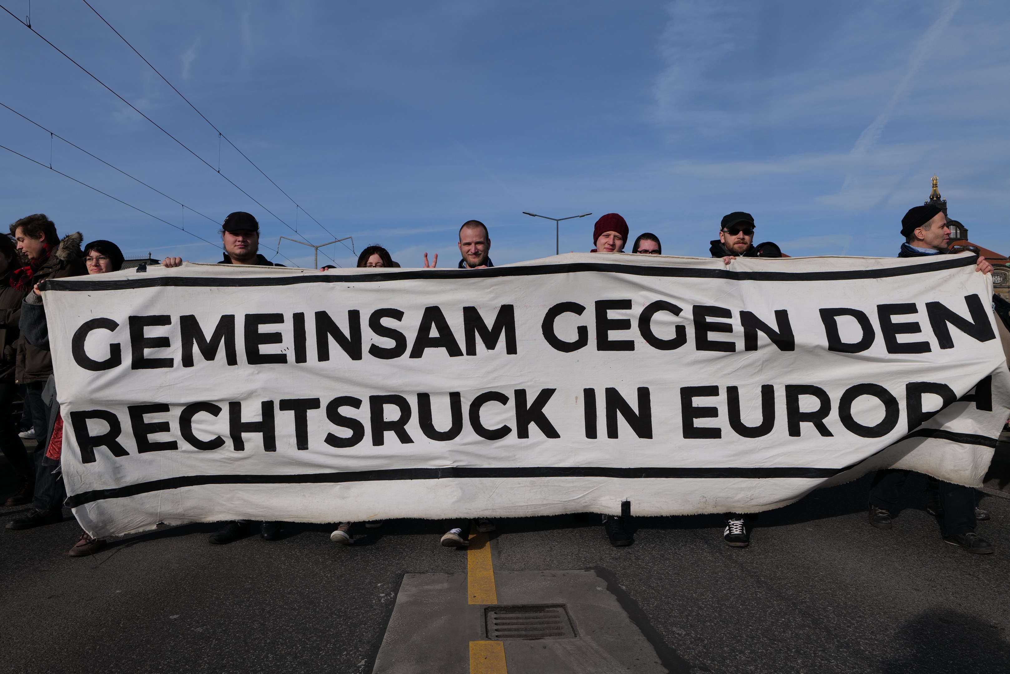 Eine Gruppe von Menschen hält ein Transparent mit der Aufschrift "Gemeinsam gegen den Rechtsruck in Europa" und protestiert gegen die Europäische Union auf einer Straße mit Straßeneinrichtungen und Gebäuden im Hintergrund.