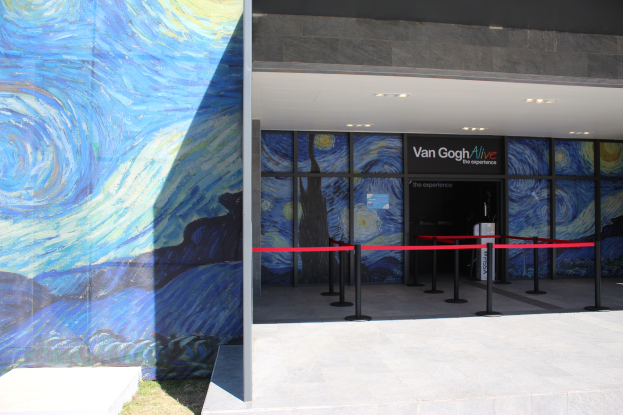 Der Eingang eines Gebäudes zeigt ein großes Wandgemälde, ein Schild mit der Aufschrift "Van Gogh Alive", Absperrpoller mit Bändern, Gras auf dem Boden und Lampen an der Decke.