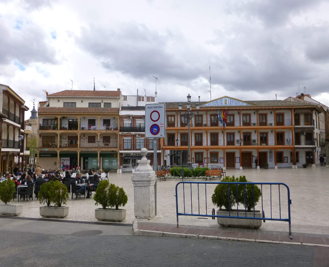 Ein belebter Stadtplatz mit Menschen, die sitzen und stehen, Topfpflanzen, Metallabsperrungen, ein Schild an einem Pfahl, Fahnen, Gebäude mit Fenstern und einem bewölkten Himmel.