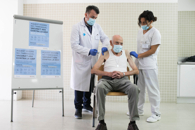 Alter Mann erhält Impfung von zwei maskierten Ärzten in einem Krankenhauszimmer, mit einer Tafel und medizinischen Geräten in der Nähe.