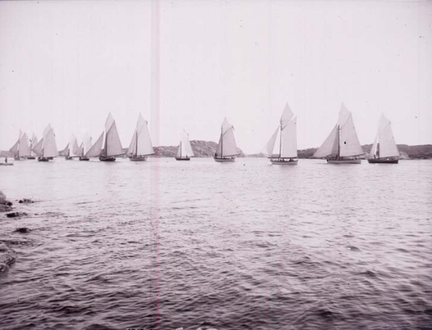 Schwarzes und weißes Foto von Segelbooten auf dem Wasser mit Bergen im Hintergrund und einem klaren Himmel, als Foto des New York Yacht Club etikettiert.