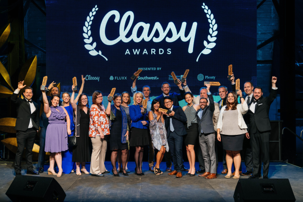 Gruppe von Menschen auf einer Bühne, die Auszeichnungen halten, mit Lautsprechern darunter und einem Bildschirm, der "Classy Awards 2019" in den Hintergrund zeigt.