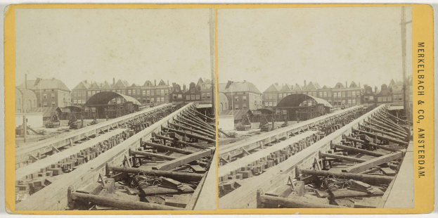 Schwarz-weißes Foto einer Bahnschiene mit Gebäuden im Hintergrund, Text 'Merkelbach & Co, Co. Amsterdam' rechts und verstreuten Holzobjekten im Vordergrund.