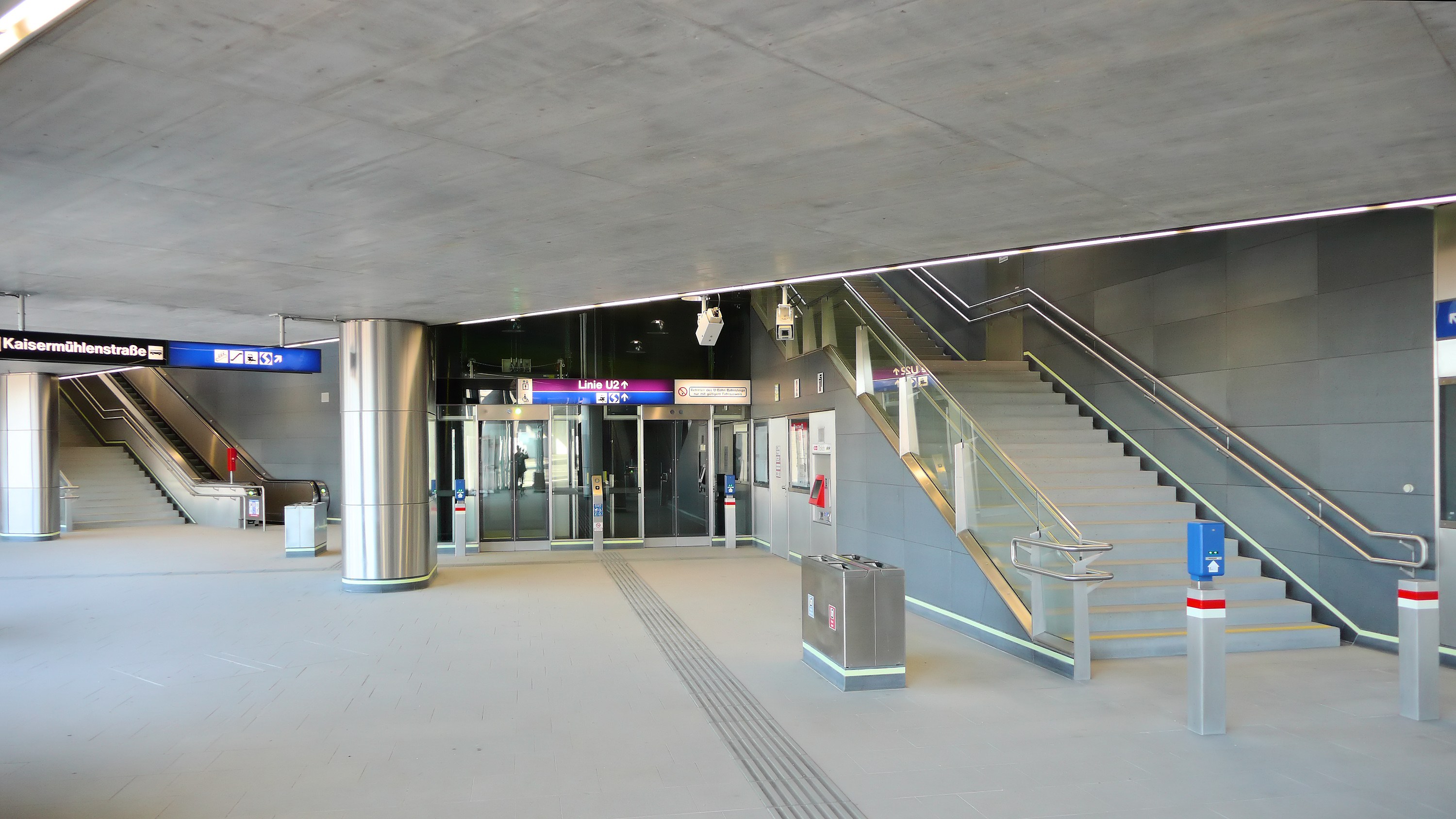 Leerer Bahnhof mit Treppe, Geländern, bodennähen Abfälleimern, Deckenleuchten, Texttafeln und Glastüren im Hintergrund.