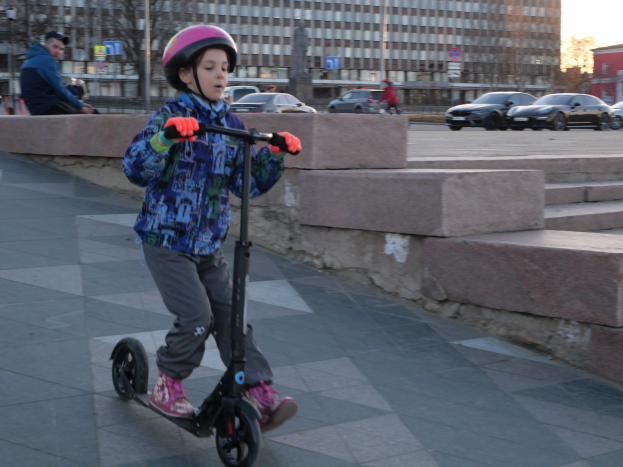 Ein junger Junge fährt mit einem Helm und Handschuhen einen Roller auf einem Gehweg, mit verschiedenen städtischen Elementen und einem klaren blauen Himmel im Hintergrund.