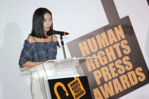Eine Frau in einem schwarzen Kleid steht an einem Rednerpult mit einem Mikrofon und hält eine Rede bei den Human Rights Press Awards, mit einem konzentrierten und ernsten Gesichtsausdruck.