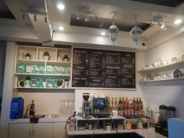 Ein gemütliches Cafe-Interieur mit einer Theke, die Flaschen, Tassen und Küchenutensilien, ein Waschbecken links und Regale im Hintergrund zeigt, die mit Geschirr und Gläsern bestückt sind, beleuchtet von Deckenlampen und einer Wandtafel.