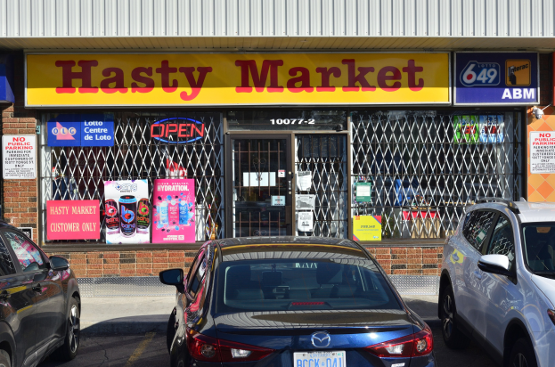 Ein geschäftiges Hasty Market-Geschäft in Toronto, Ontario, mit Autos auf der Straße vor dem Geschäft und Glaswänden mit Postern.
