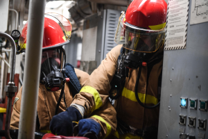 Zwei Feuerwehrleute in Schutzausrüstung arbeiten an einem Feuerwehrauto, mit einer Tafel und Metallstangen im Hintergrund.