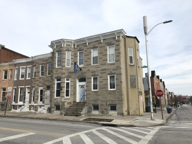 Eine Reihe moderner Häuser an einer Straßenecke in Philadelphia, mit Fenstern, Türen, Geländern, Laternen, Schildern und Fahrzeugen, sowie Bäumen und einem bewölkten Himmel im Hintergrund.