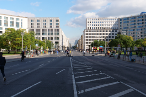 Eine belebte Stadtstraße in Berlin, Deutschland, mit Fußgängern und Radfahrern, hohen Gebäuden, Bäumen, Laternen und Schildern unter einem bewölkten Himmel.
