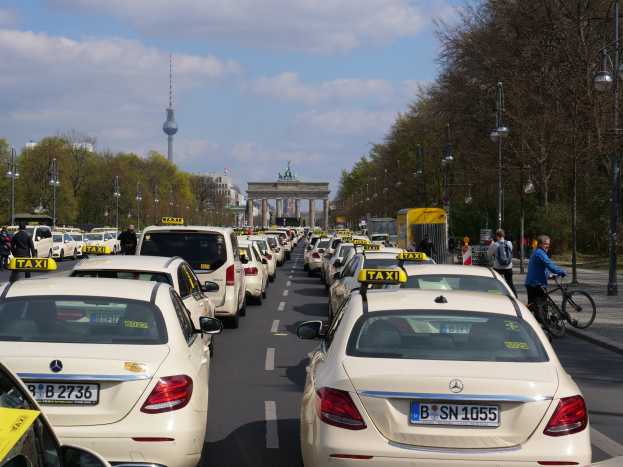 Eine lange Reihe von Taxis, die an einer belebten Straße in Berlin, Deutschland, geparkt sind, mit Fahrzeugen, Radfahrern und Fußgängern, flankiert von Laternenpfählen, Bäumen und Gebäuden, einschließlich eines Bogens und eines Turms, unter einem bewölkten Himmel.