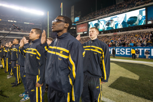 Eine Gruppe von Männern in blauen und gelben Jacken steht auf einem Fuballfeld, mit einer Menge, Schildern, Fahnen, Stadionbeleuchtung und Mikrofonen für eine Marschkapelle im Hintergrund.