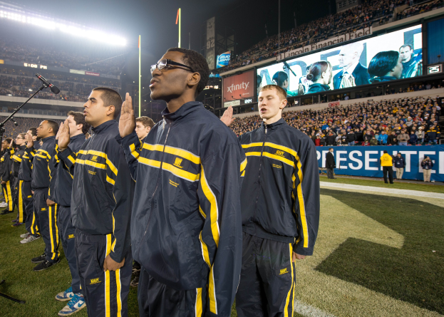 Eine Gruppe von Männern in blauen und gelben Jacken steht auf einem Fuballfeld, mit einer Menge, Schildern, Fahnen, Stadionbeleuchtung und Mikrofonen für eine Marschkapelle im Hintergrund.