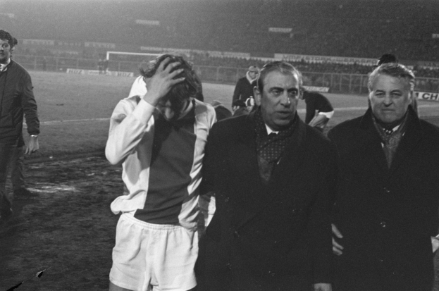 Eine Gruppe von Männern steht zusammen auf einem Fußballfeld und sieht besorgt aus, mit einem Stadion im Hintergrund, dargestellt in Schwarz-Weiß.