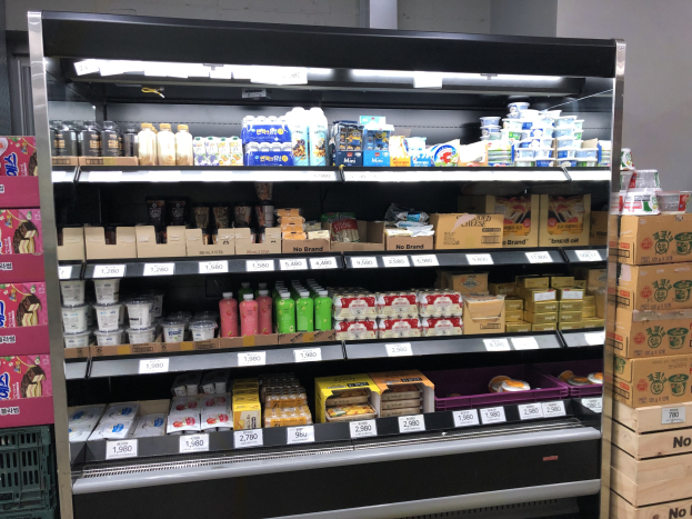 Supermarktvitrine mit verschiedenen Lebensmitteln, Flaschen, Schachteln und Preisschildern, Kartons rechts und eine Wand im Hintergrund.