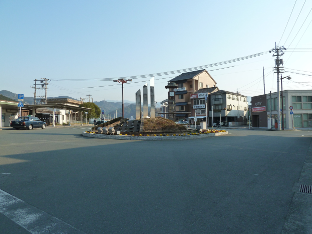A roundabout in an urban area with parked cars, buildings, electric poles, signboards, trees, distant hills, and a clear blue sky.