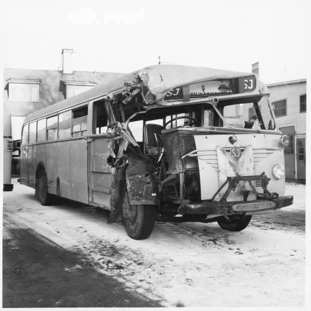 Schwarzes und weißes Foto eines schwer beschädigten Busses mit eingedellter Front und zersplitterten Fenstern am Straßenrand, mit Gebäuden im Hintergrund.