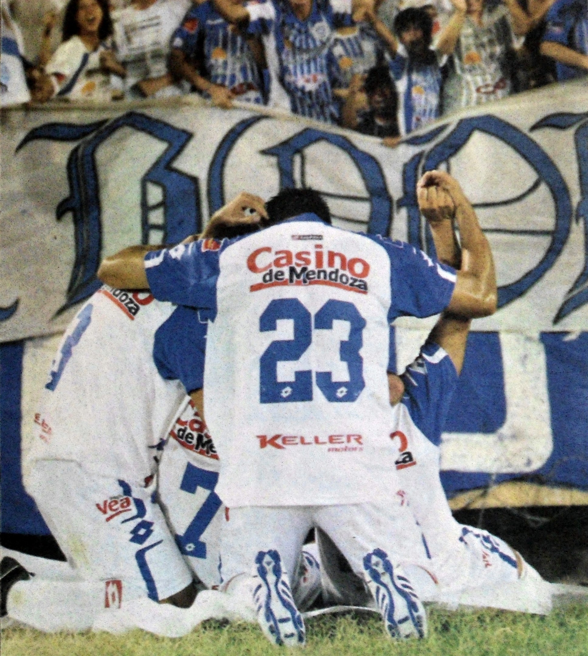 Gruppe von Füßballspielern feiert auf dem Boden vor einer jubelnden Menge mit einem Banner im Hintergrund.