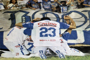 Gruppe von Füßballspielern feiert auf dem Boden vor einer jubelnden Menge mit einem Banner im Hintergrund.