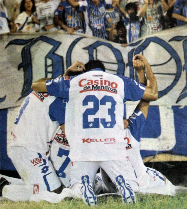 Gruppe von Füßballspielern feiert auf dem Boden vor einer jubelnden Menge mit einem Banner im Hintergrund.