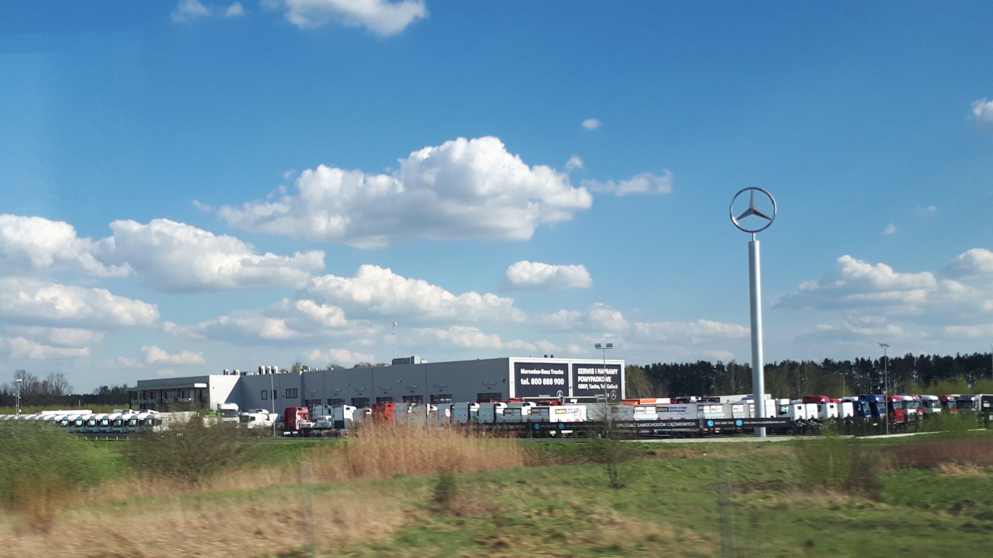 Eine Mercedes-Benz-Fabrik in Deutschland mit Fahrzeugen auf der Straße, Grünflächen, Gebäuden und einem logo-beschrifteten Pfahl unter einem bewölkten Himmel.