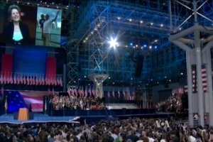 Eine große Menschenmenge sitzt vor einer Bühne mit einem Podium, Mikrofon, Flaggen, einem Bildschirm und Lichtausrüstung und wartet auf eine Präsidentschaftsdiskussion.