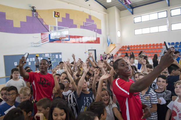 Gruppe von Kindern vor einem Basketballfeld mit mobilen Telefonen, einer Anschlagtafel, einer Uhr, einem Torpfosten, einem Basketballkorb, Deckenleuchten, Stühlen und Fenstern im Hintergrund.