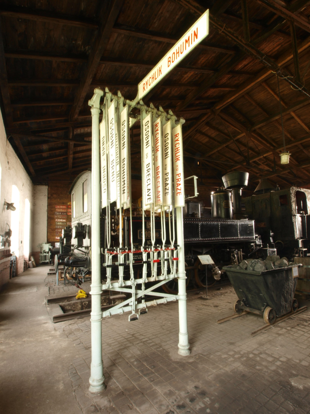 Ein Bahnhof mit einem Zug auf den Gleisen, einem Rollstuhl auf dem Boden, einem Pfahl mit einer Tafel, einer Wand auf der linken Seite, einer Decke oben und einer zentralen Signalbox, die den Zugang der Züge steuert.
