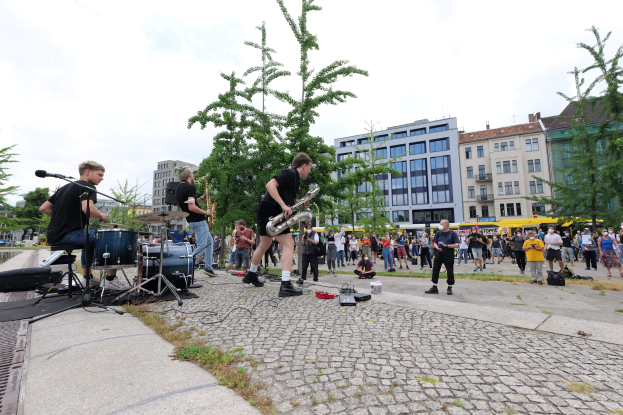 Eine Gruppe von Menschen, die Musik auf einem Bürgersteig mit verstreuter Ausrüstung spielt, vor einem Publikum, mit Bäumen, Gebäuden und einem bewölkten Himmel im Hintergrund.