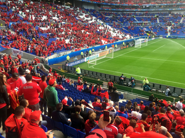 Eine große Menschenmenge in einem Stadion schaut ein Fußballspiel zu, mit Menschen auf Stühlen und Stehplätzen, Bannern, einem Torpfosten und Deckenbeleuchtung.