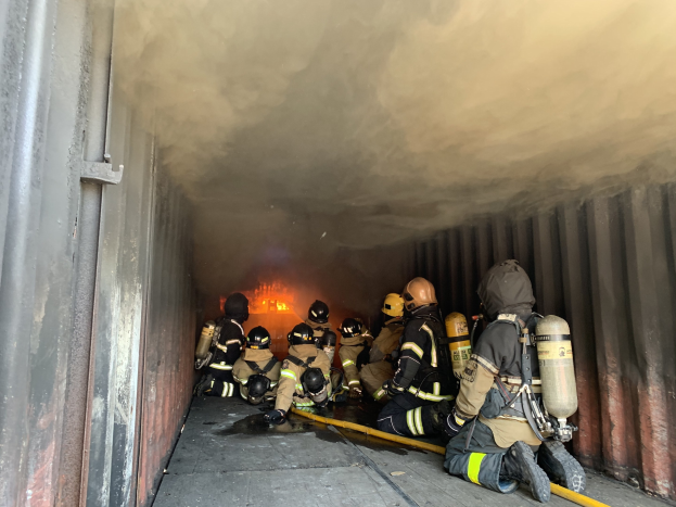 Feuerwehrleute mit Helmen und Sauerstoffflaschen arbeiten in einem raucherfüllten Tunnel mit einem sichtbaren Feuer im Hintergrund, einer hält ein Rohr und eine Wand ist links zu sehen.