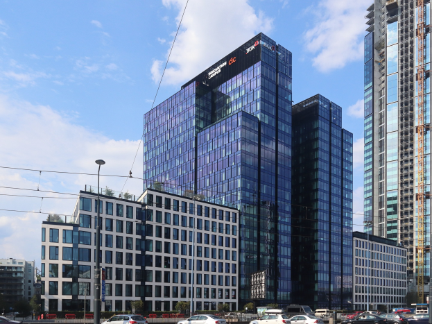 Zentrale der Deutschen Bank in Frankfurt, Deutschland, ein großes mehrstöckiges Gebäude mit zahlreichen Fenstern, umgeben von städtischer Infrastruktur einschließlich Straßenlaternen, Stromkabeln, Fahrzeugen, Grünflächen und einem bewölkten Himmel.