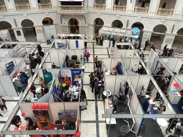Eine große Gruppe von Menschen auf einer internationalen Fachmesse mit Ständen, Bannern, Tischen, Stühlen, Luftballons und einem Gebäude mit Bögen, Säulen, Geländern und einem Bildschirm im Hintergrund.