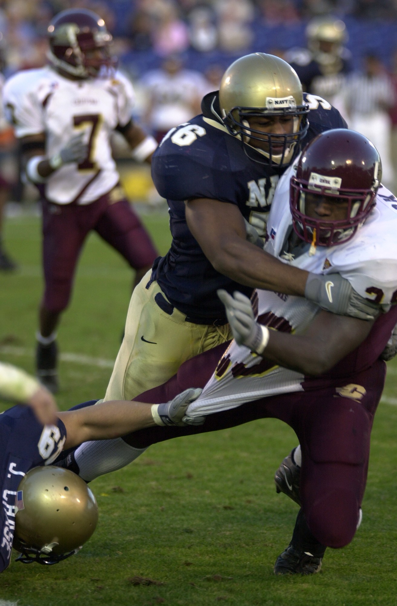 Ein Fussballspieler in einem Helm wird von einem anderen Spieler auf dem Feld getackelt, mit anderen Spielern und Zuschauern im Hintergrund.
