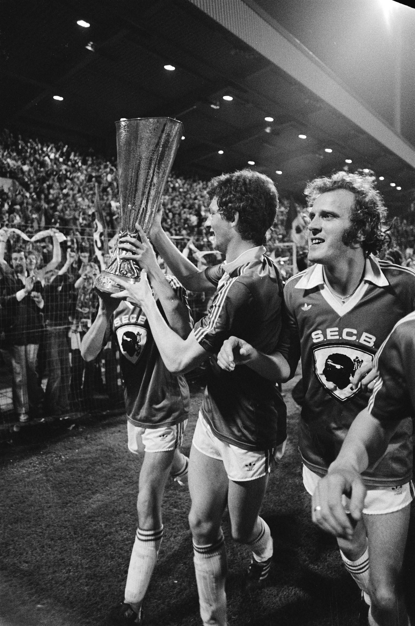 Schwarzes und weißes Bild von Fußballspielern, die einen Pokal auf einem Feld halten und ihren FA-Cup-Finalsieg feiern, während Zuschauer hinter einem Zaun zusehen.