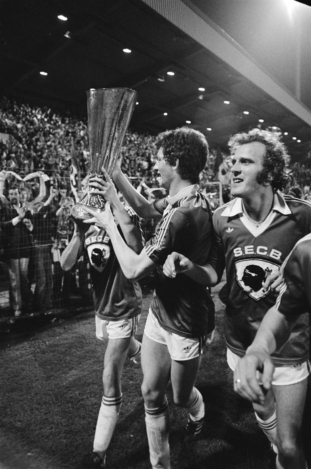 Schwarzes und weißes Bild von Fußballspielern, die einen Pokal auf einem Feld halten und ihren FA-Cup-Finalsieg feiern, während Zuschauer hinter einem Zaun zusehen.