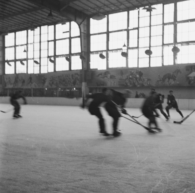 Schwarzes und weißes Foto von Menschen, die Hockey auf einem Eisplatz spielen, mit einer bemalten Wand und Glasfenstern im Hintergrund.