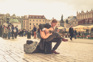 Ein Mann sitzt auf dem Boden und spielt Gitarre in einem belebten Stadtplatz, umgeben von Passanten, mit einer Tasche auf dem Boden und Gebäuden, einer Statue und einem klaren Himmel im Hintergrund.