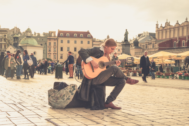 Ein Mann sitzt auf dem Boden und spielt Gitarre in einem belebten Stadtplatz, umgeben von Passanten, mit einer Tasche auf dem Boden und Gebäuden, einer Statue und einem klaren Himmel im Hintergrund.