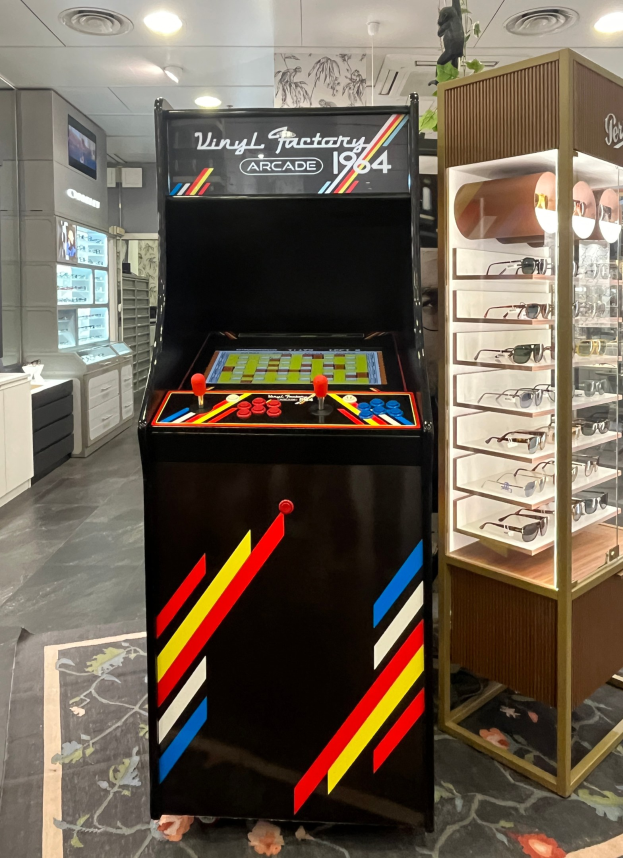 Ein Geschäft mit einem zentralen Retro-Arcade-Automaten, einem rechten Gestell mit Gläsern und Hintergrund-Elementen wie Schränken, einem Fernseher und anderen Gegenständen, mit Deckenlampen, die eine Retro-Atmosphäre schaffen.