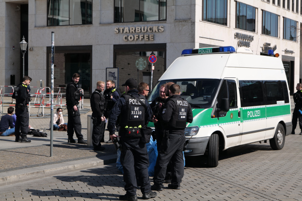 Polizisten vor einem Starbucks-Café mit einem Van auf der rechten Seite und Menschen auf der linken Seite, vor einer Gebäudefassade, einem Schild, einem Laternenpfahl und einem Zaun.
