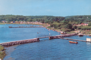 Ein altes Foto einer See-Szene mit einem Boot, einer Brücke, Flaggen, Felsen, Bäumen, Gebäuden und einem Himmel, das viele Menschen beim Schwimmen zeigt und Text auf dem Bild.