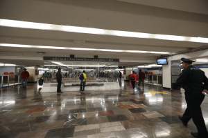 Eine Gruppe von Menschen geht durch eine belebte U-Bahn-Station, mit einem Polizisten im Vordergrund, umgeben von Geländern, Bildschirmen und Texttafeln an der Decke.